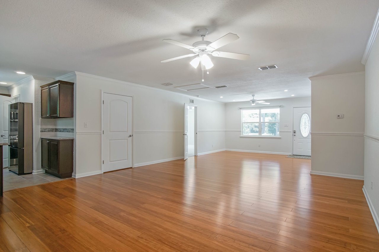 Spacious empty living room with wooden floors, ceiling fan, and recessed lighting. Sunlight streams through a window next to a white front door.