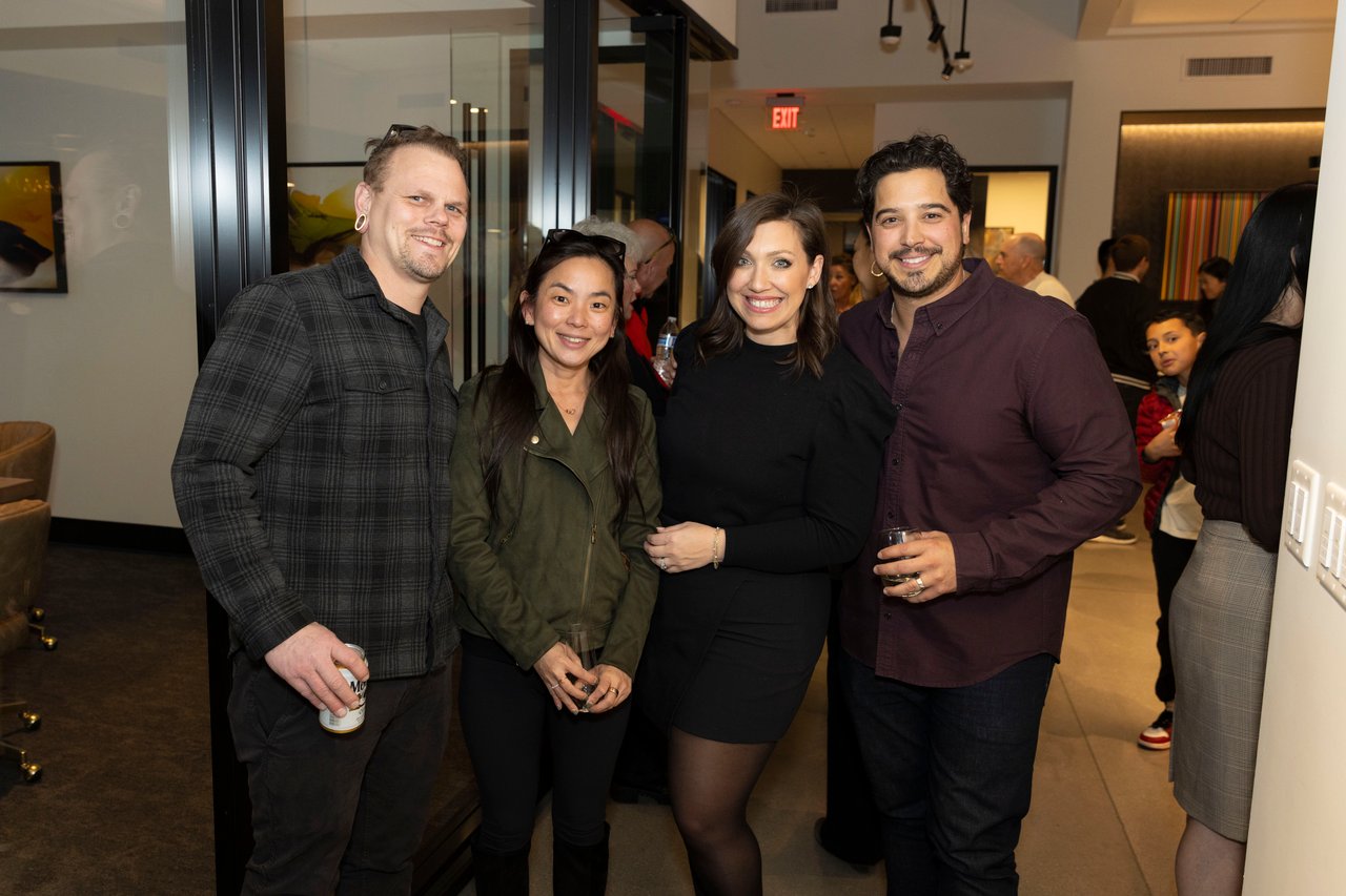 Four guests pose together at an indoor event, with other attendees in the background.