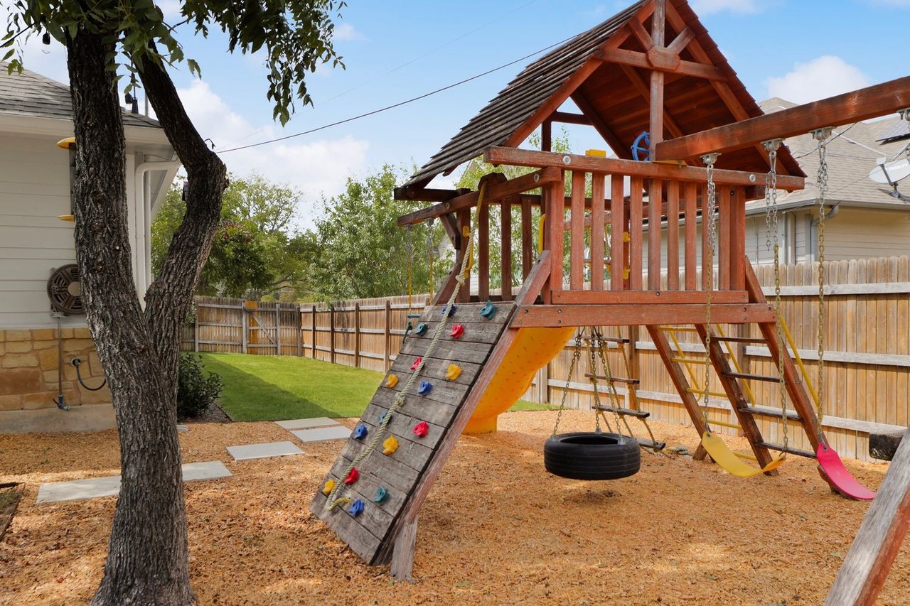Fenced backyard with a large wooden playset, colorful climbing holds, swings, and slides.