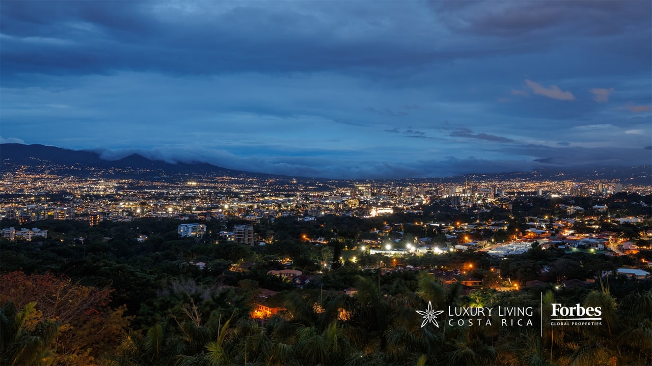Twilight city skyline beneath cloud-covered mountains, with lights spread across the valley.
