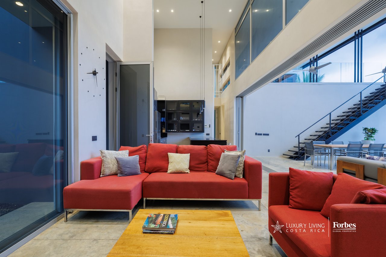 Double-height living room with red sectional sofas, floor-to-ceiling glass walls, and an open staircase.