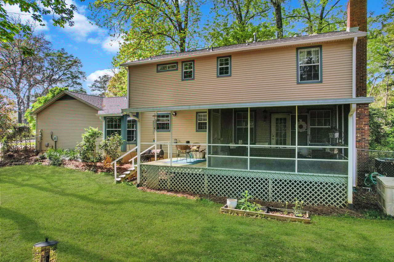 An exterior view of a two-story house with a well-maintained lawn. The house has a small porch and is surrounded by trees and greenery.