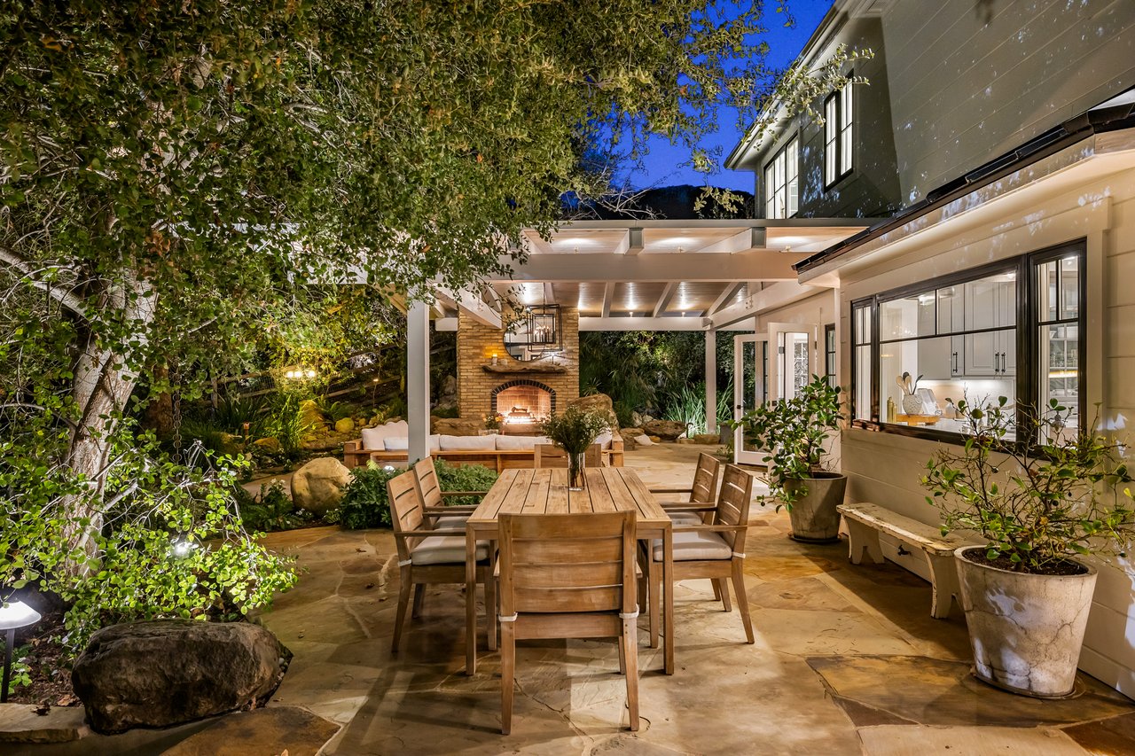 Outdoor dining patio with a pergola, garden landscaping, and lit brick fireplace beside the house.