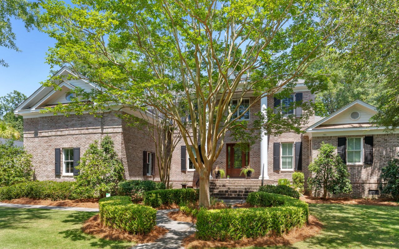 Brick home with white trim and dark shutters, shaded by a large tree over the landscaped front lawn.