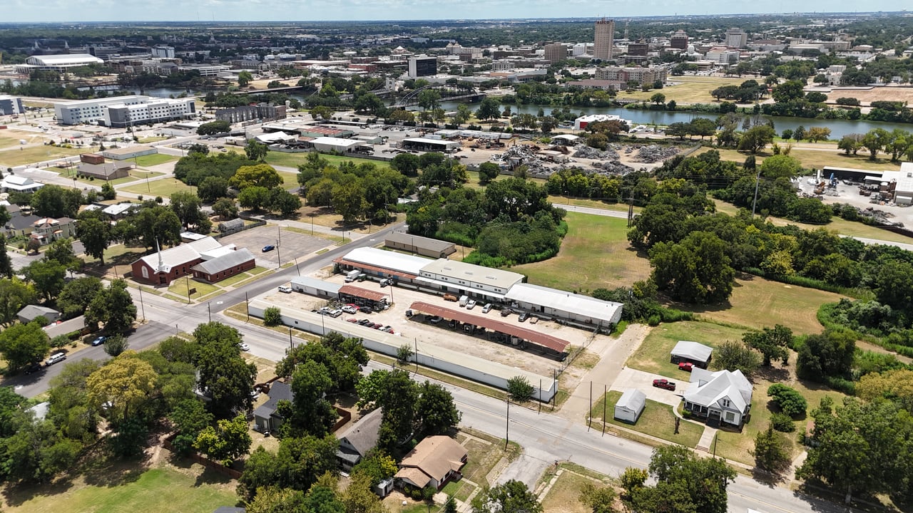 Aerial view of commercial warehouses and surrounding grounds, with a river and city skyline beyond.