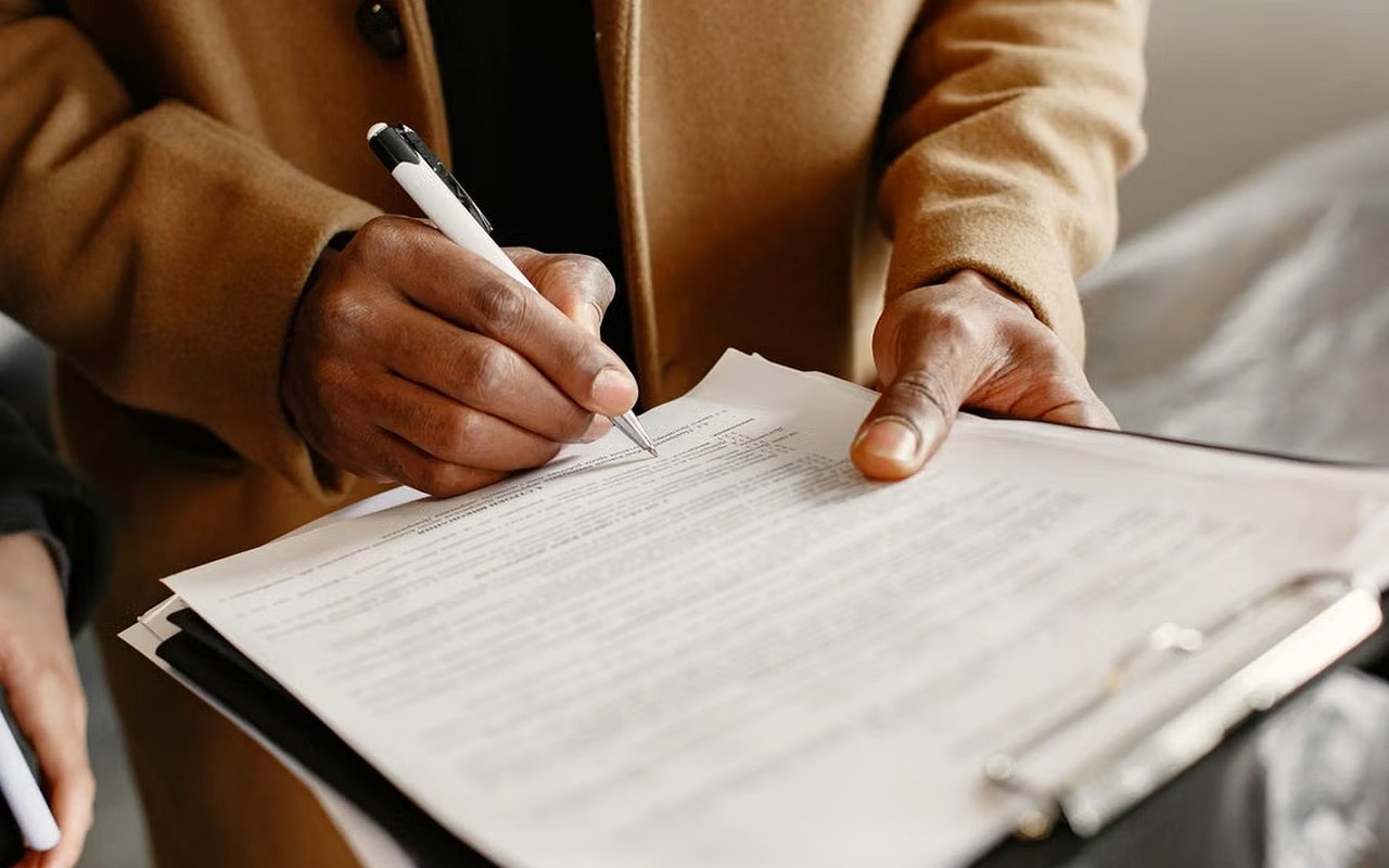 A person signs a document on a clipboard with a pen.