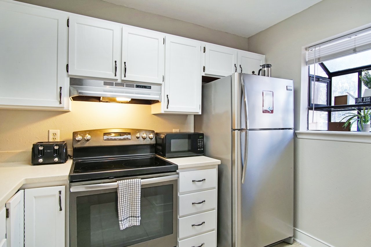 Compact kitchen with white cabinets, stainless steel fridge, and oven. A checkered towel hangs on the oven. Bright window light creates a welcoming feel.