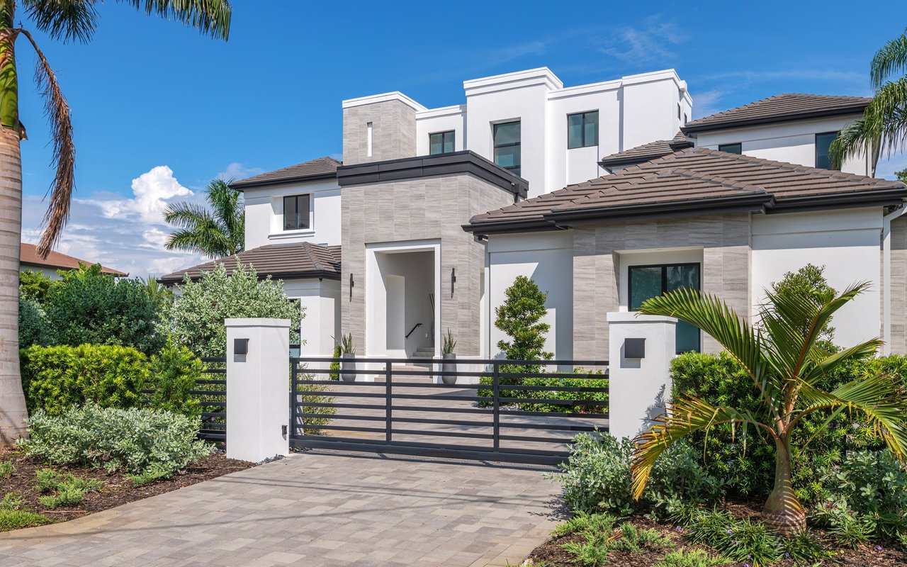 Modern white home behind a black metal gate, framed by palm trees and tropical landscaping.