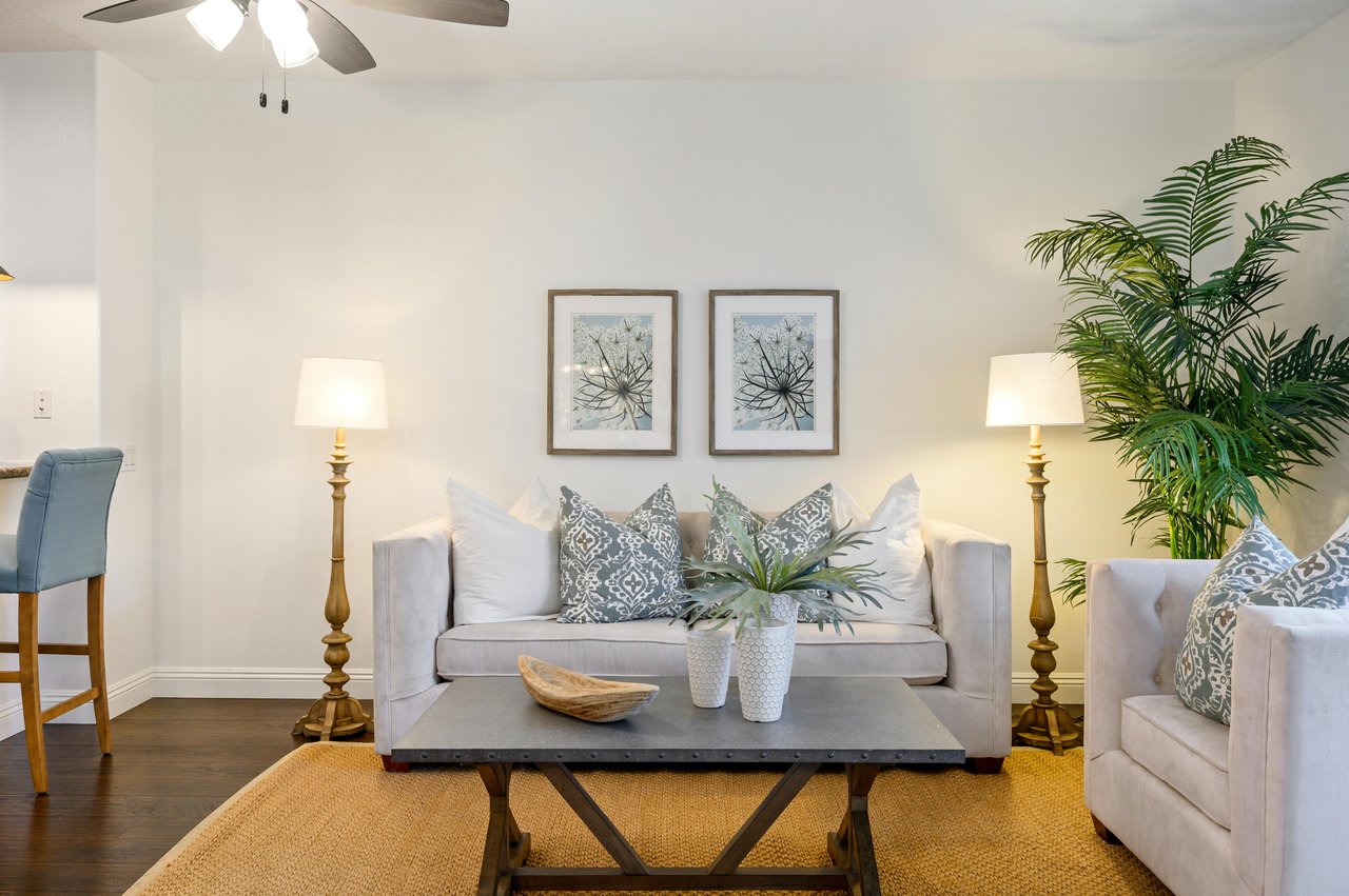 Bright living room with white sofas, patterned pillows, a coffee table, tall lamps, and framed wall art.