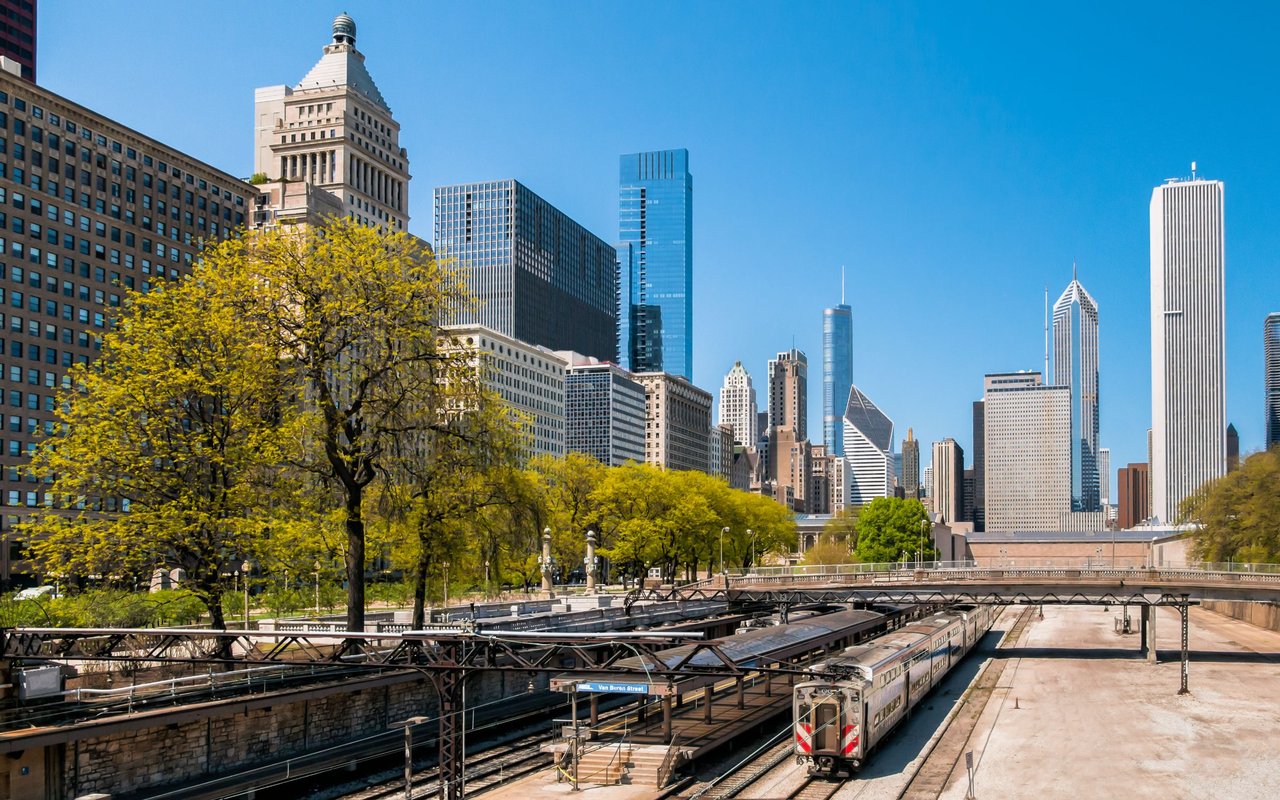 Chicago skyline above elevated train tracks, with a CTA train and autumn-colored trees.