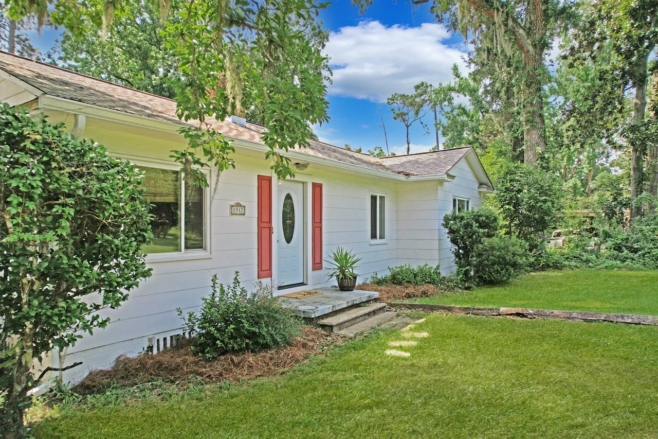 Small white house with a red door, surrounded by lush green plants and trees. Bright blue sky and peaceful, inviting atmosphere.