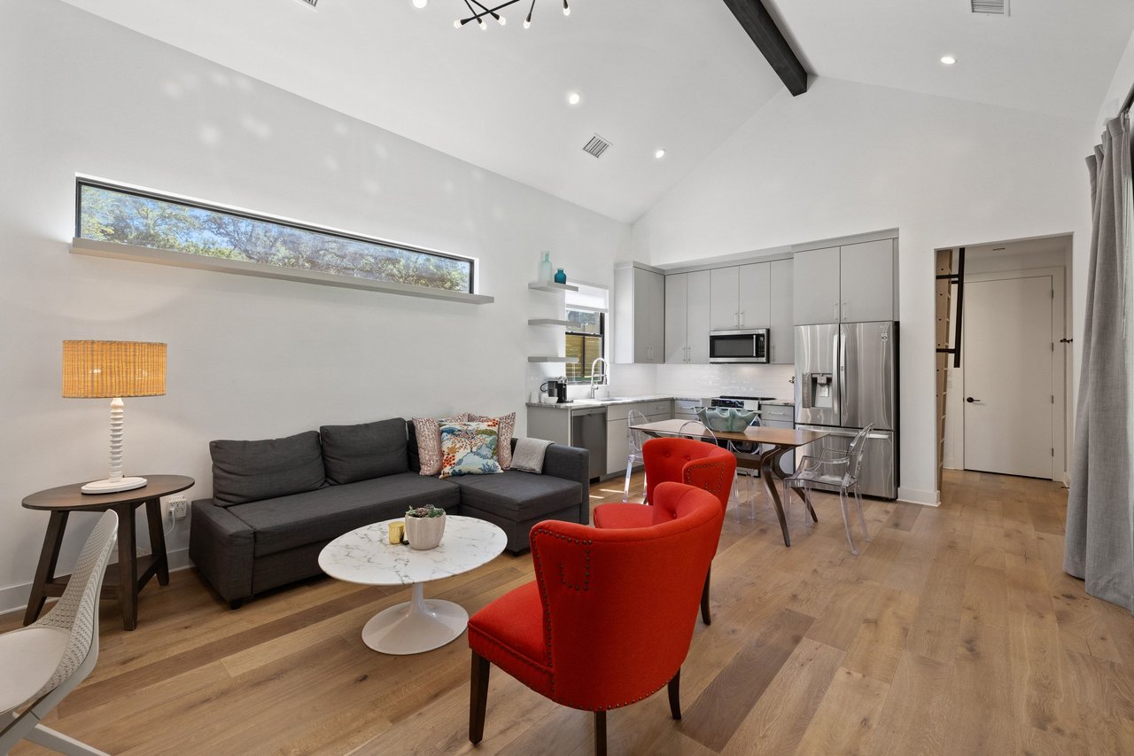 Open-plan living room and kitchen with a gray sectional, red chairs, stainless steel appliances, and vaulted ceiling.