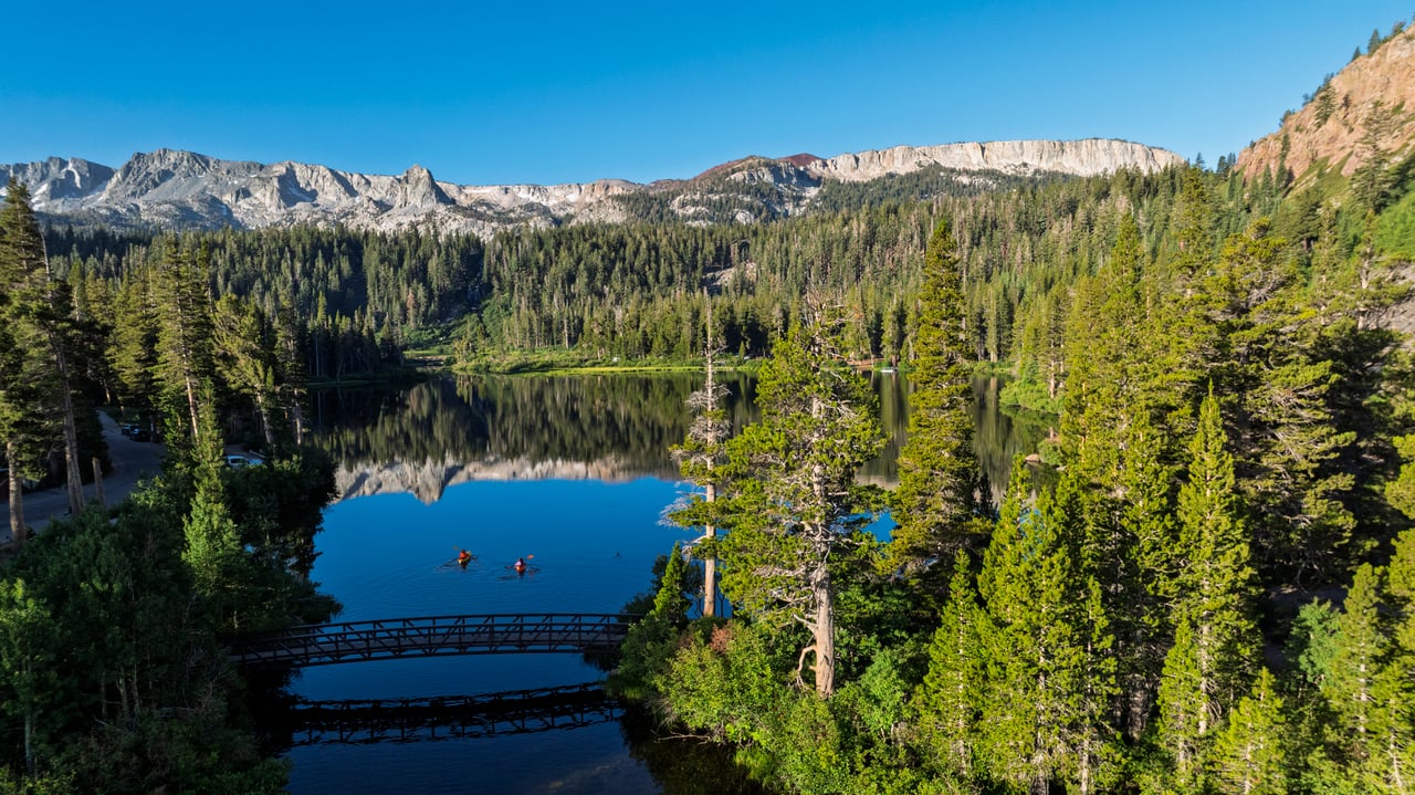 Forest-fringed mountain lake reflecting rocky peaks, with a footbridge and two kayakers.
