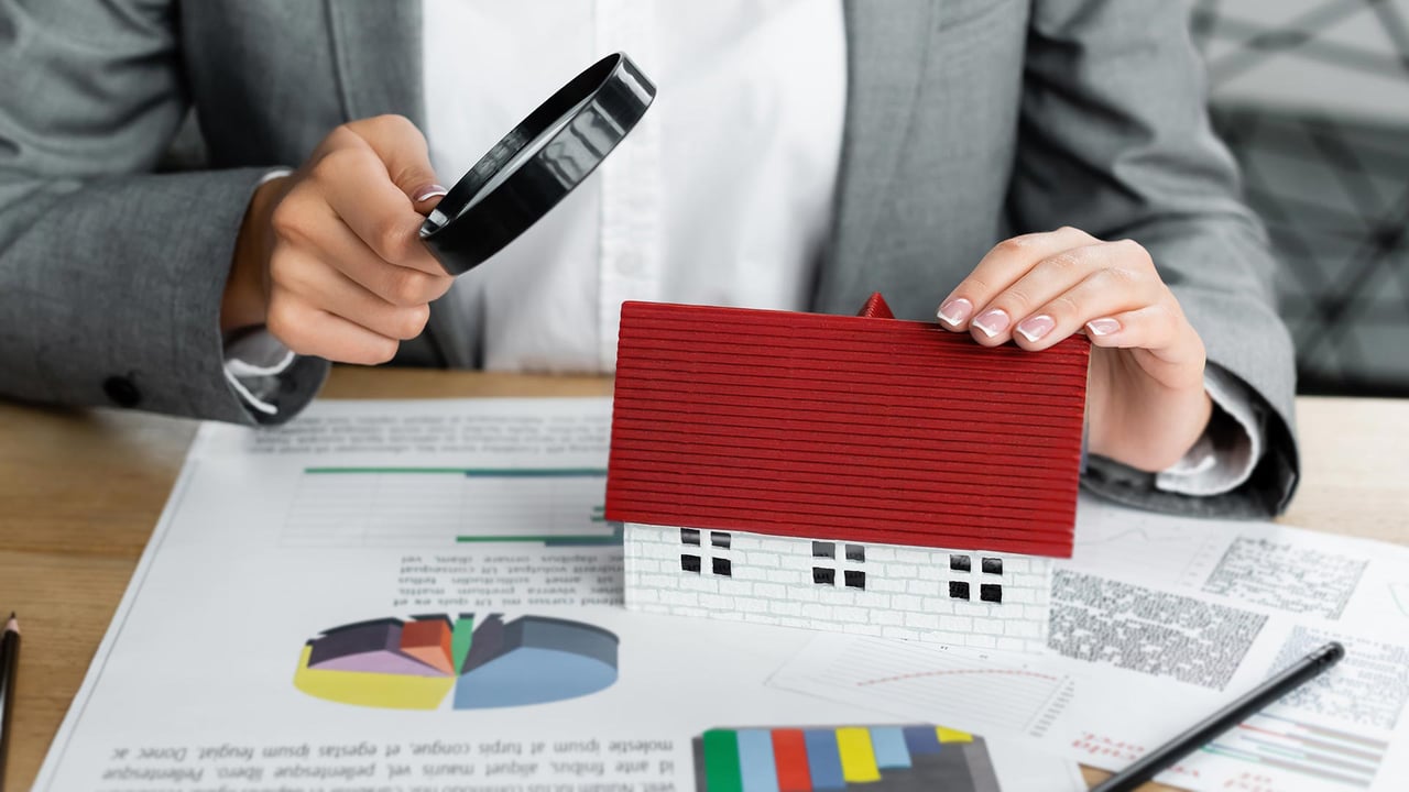 A close up of a professional holding a magnifying glass over a small model house on a desk covered in real estate market charts, representing a meticulous human analysis for a precise free home evaluation in Mississauga and Oakville