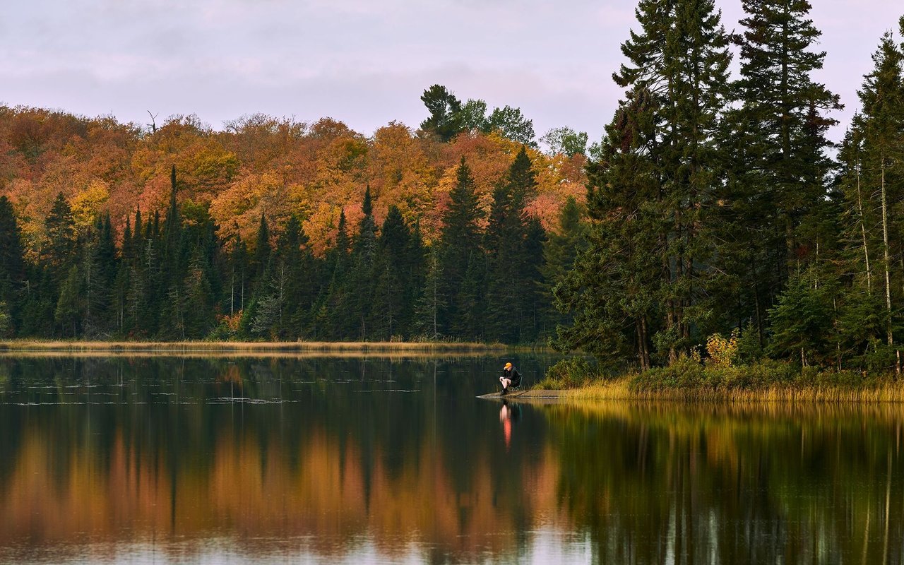 Autumn lake reflecting colorful woodland, with a lone angler at the water’s edge.