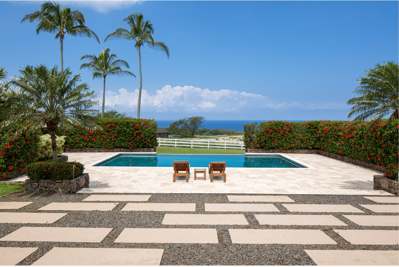 Swimming pool and stone patio framed by palms and flowering hedges, with ocean beyond.
