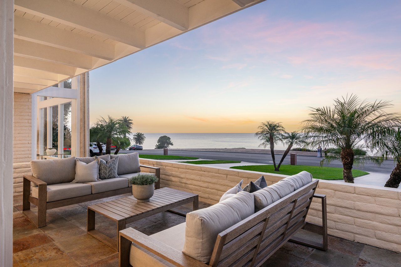 Furnished patio overlooking the ocean at sunset, with cushioned seating and palm trees.