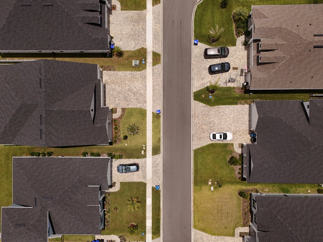 Aerial view of homes along a residential street, with paved driveways, parked cars, and lawns.