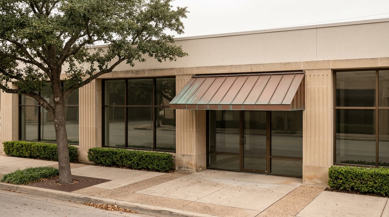 Historic storefront with fluted stone piers, glass doors, a copper awning, and a live oak shading the sidewalk.