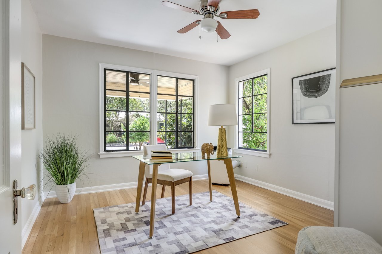 Staged home office with a glass-top desk, geometric rug, and black-grid windows overlooking greenery.