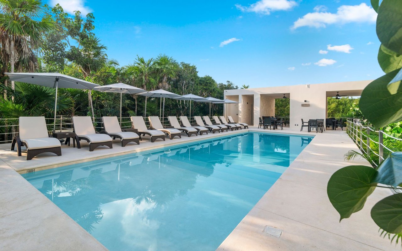 Outdoor pool with rows of lounge chairs and umbrellas beside a covered pavilion and tropical landscaping.