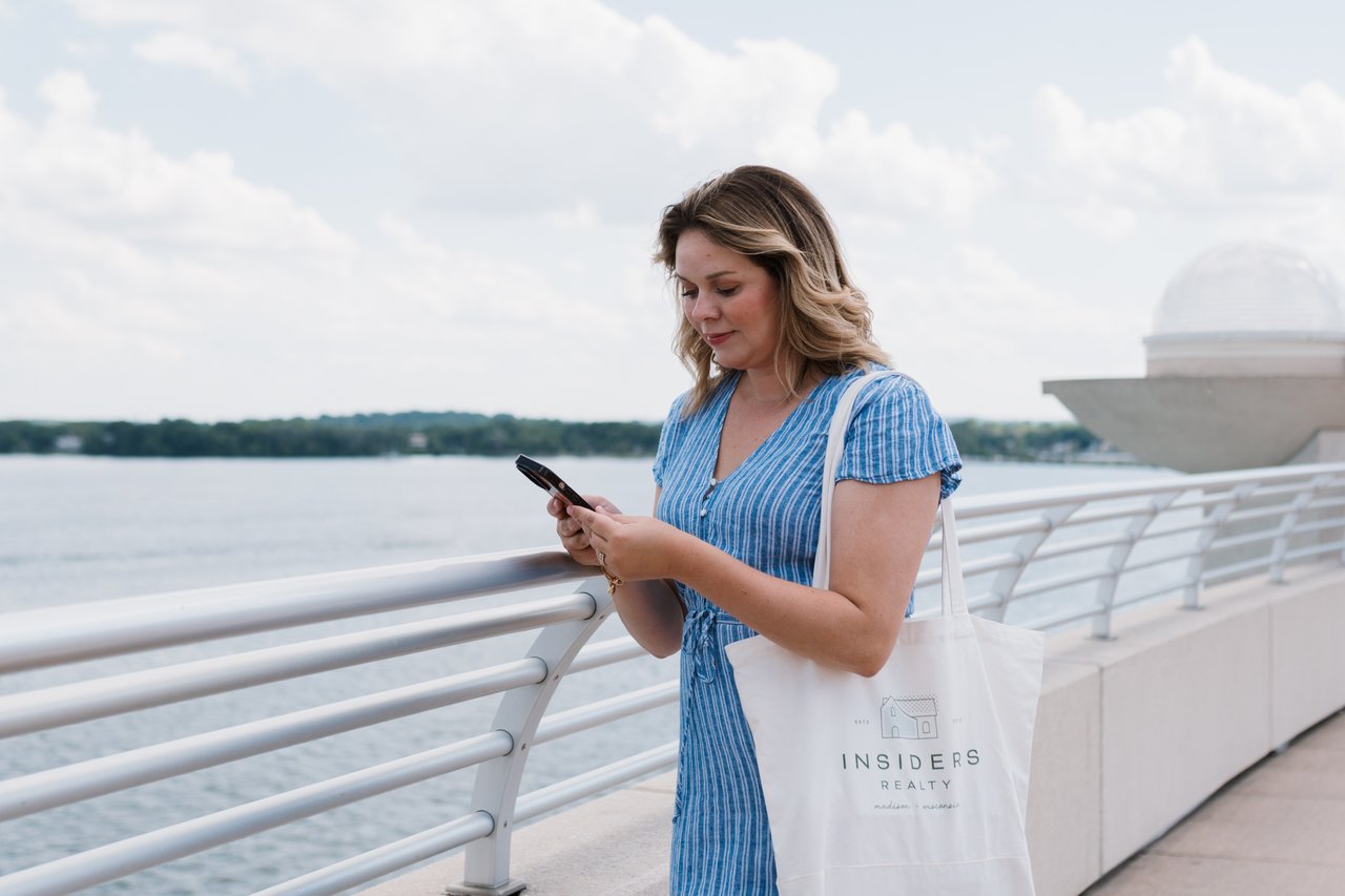 Cait Berry of Insiders Realty standing at Monona Terrace overlooking Lake Monona on a summer day, checking her phone while carrying an Insiders Realty tote, with the John Nolen Drive corridor visible along the south shore.