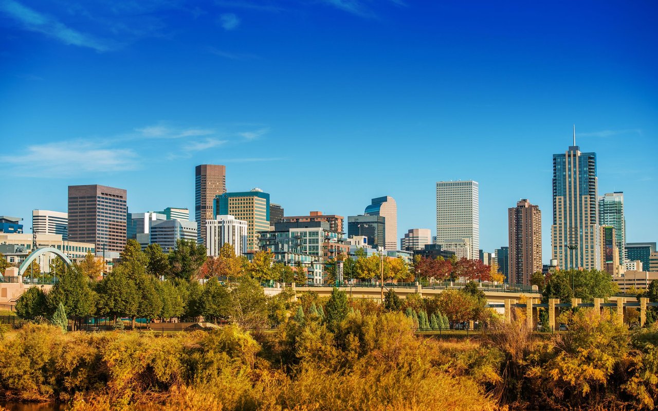 Denver skyline beyond a bridge and autumn-colored trees.