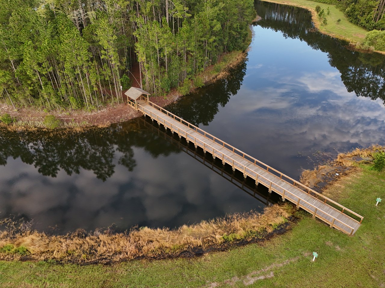 A pond bordered by trees, with a long wooden boardwalk leading to a covered pavilion.