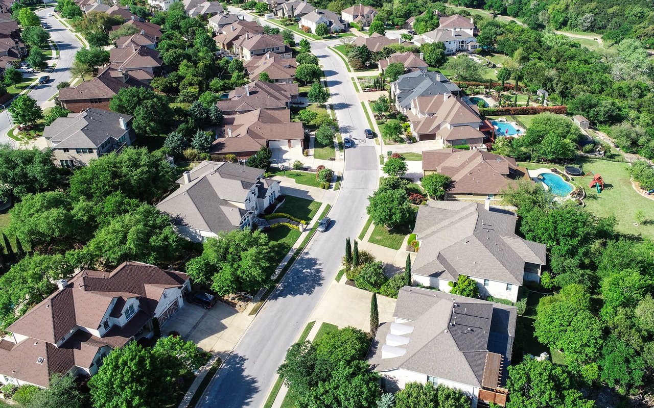 Aerial view of a leafy neighborhood with detached homes, curving streets, and backyard pools.