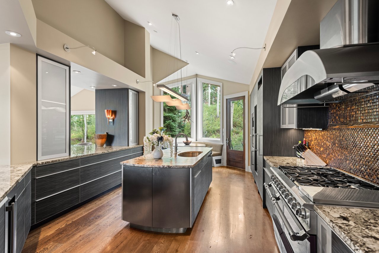 Contemporary kitchen with a stone island, dark cabinetry, professional range, and tall windows overlooking trees.