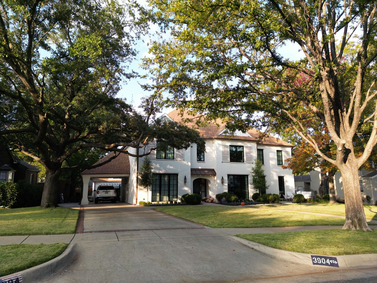 White two-story house framed by mature trees, with a covered carport and broad driveway.