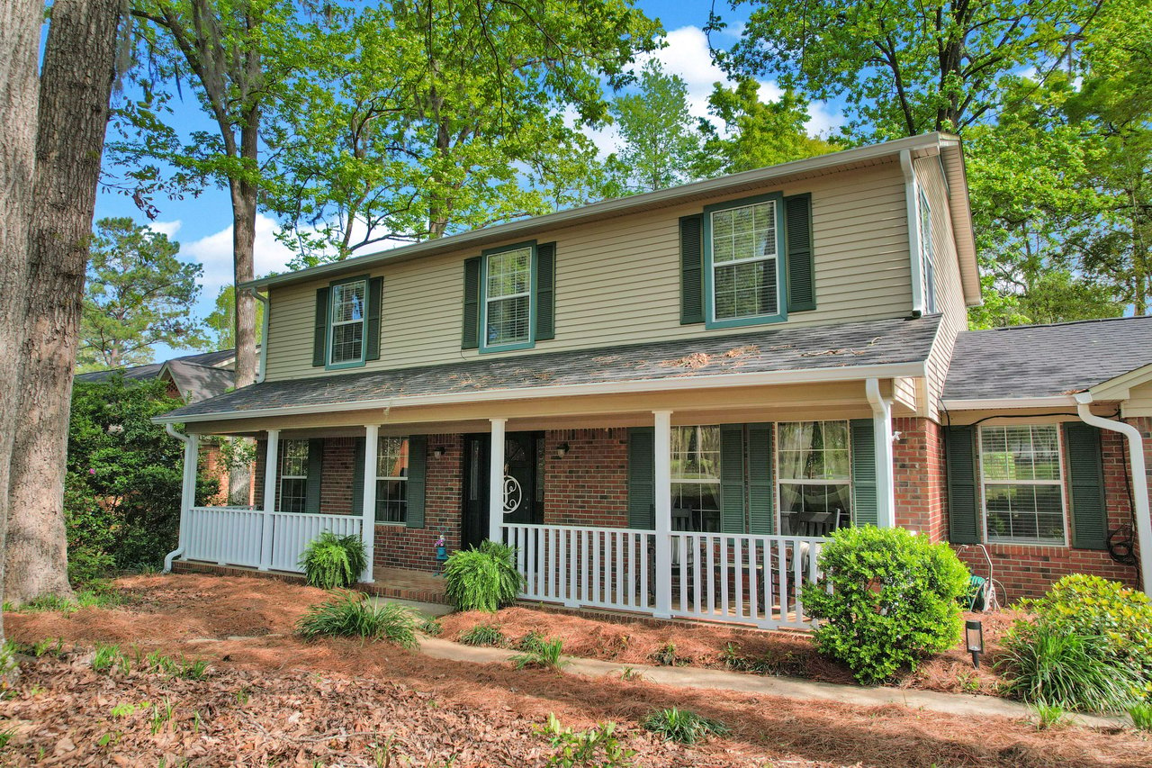 An exterior view of a two-story house with a large front porch. The house is surrounded by well-maintained landscaping and tall trees.