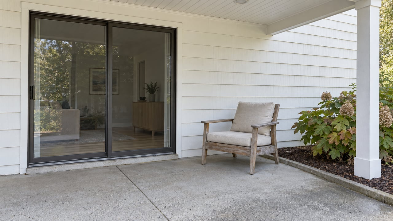 A covered patio with white siding, closed sliding glass doors, a weathered cedar chair, and an oakleaf hydrangea in a curbed bed.