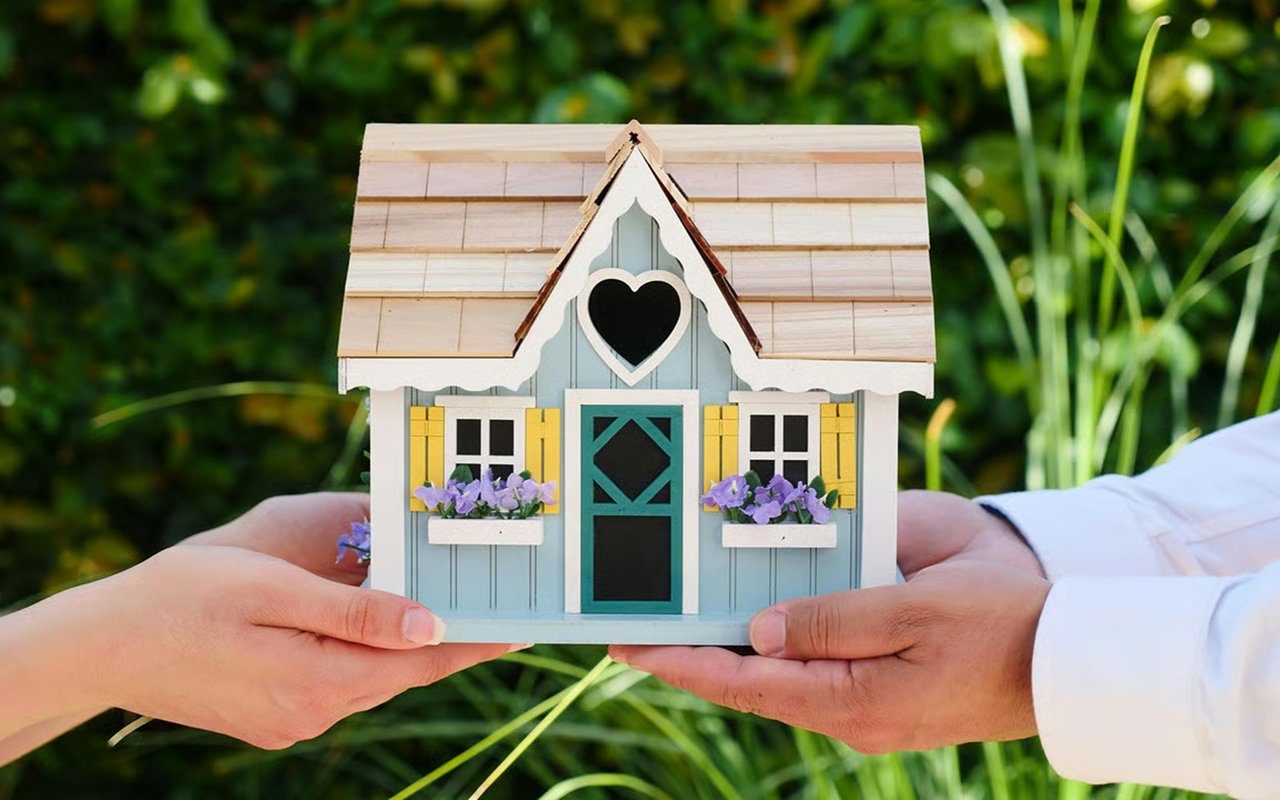 Hands hold a miniature blue house with yellow shutters, purple flowers, and a heart-shaped opening.