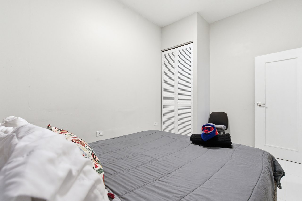 Bedroom with a gray quilted bed, louvered closet doors, and folded towels on a chair.