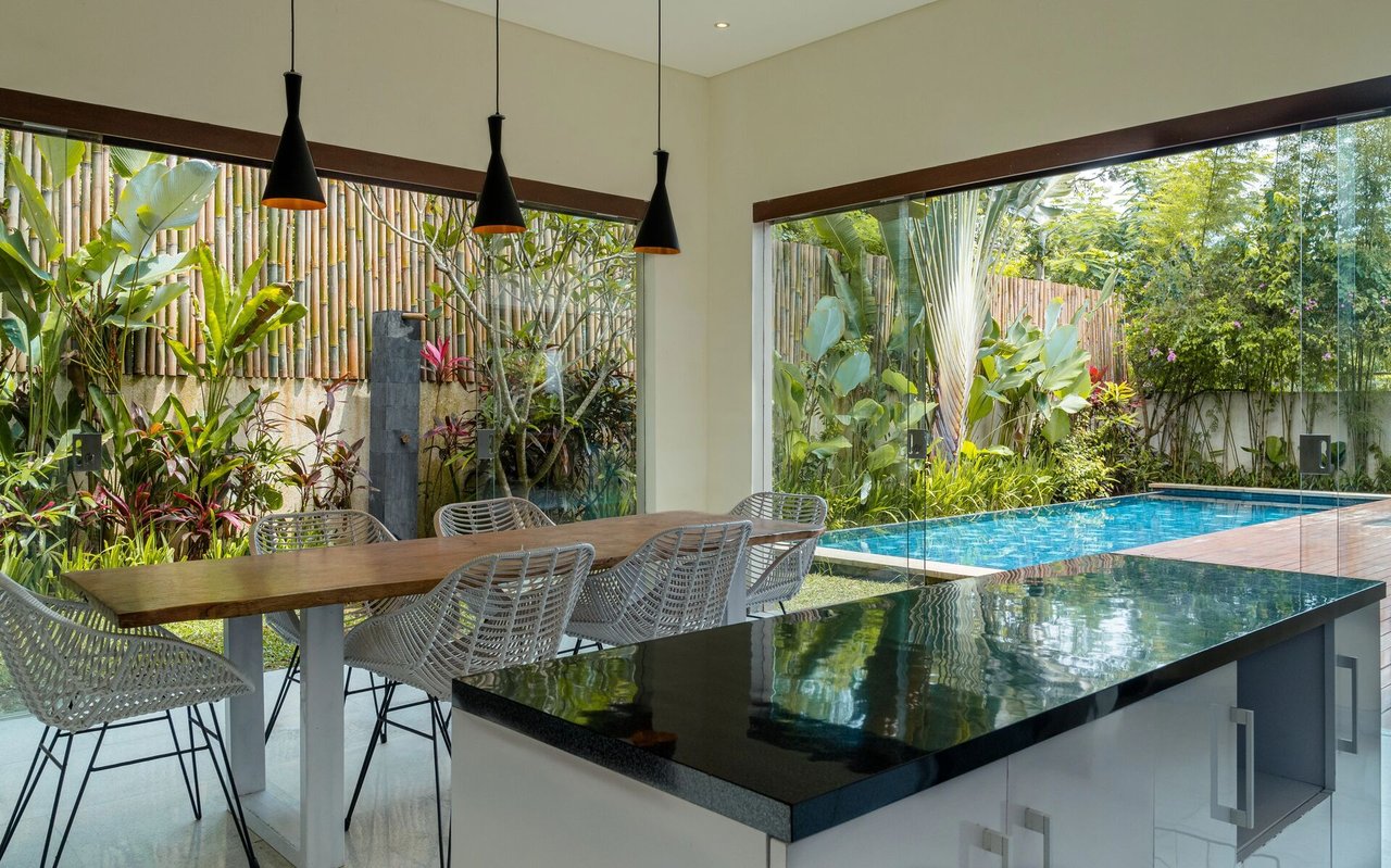 Dining area with woven chairs and a kitchen island overlooking a tropical garden and pool.