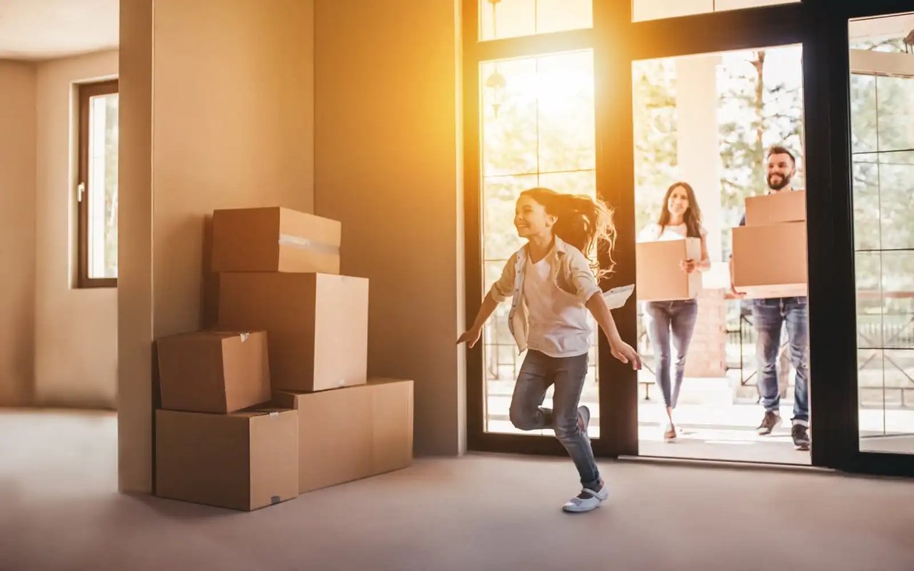 A woman celebrates as a couple carry moving boxes into a sunlit home.