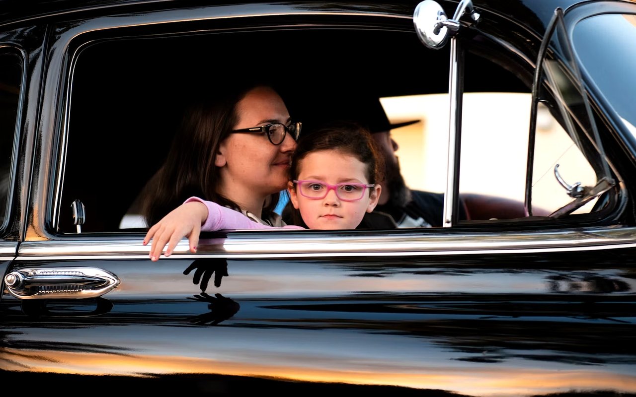 A woman and child sit in a vintage car, with the child leaning on the open window.