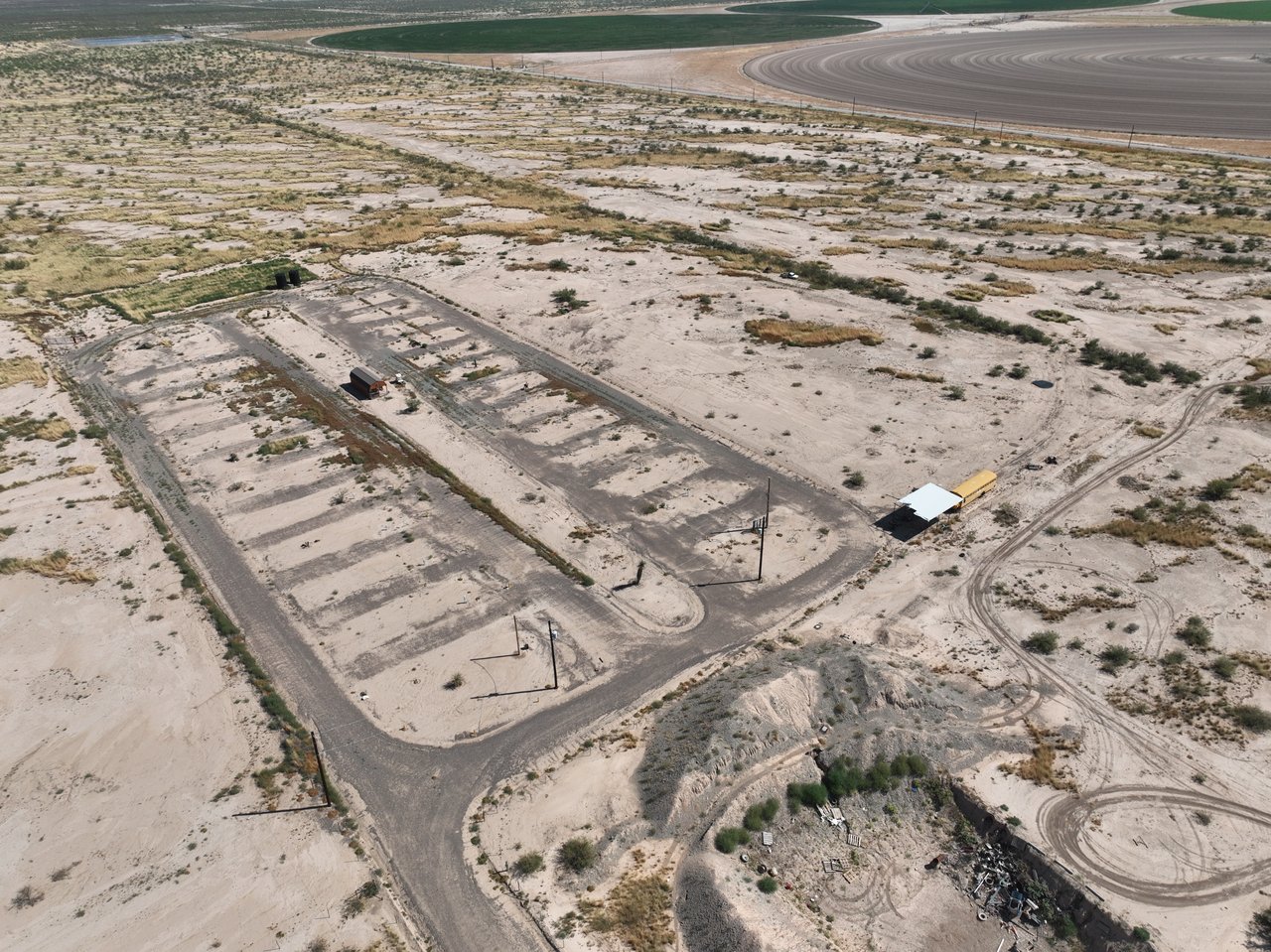 Aerial view of a sandy property with paved roads, cleared pads, and a small white-roofed structure.