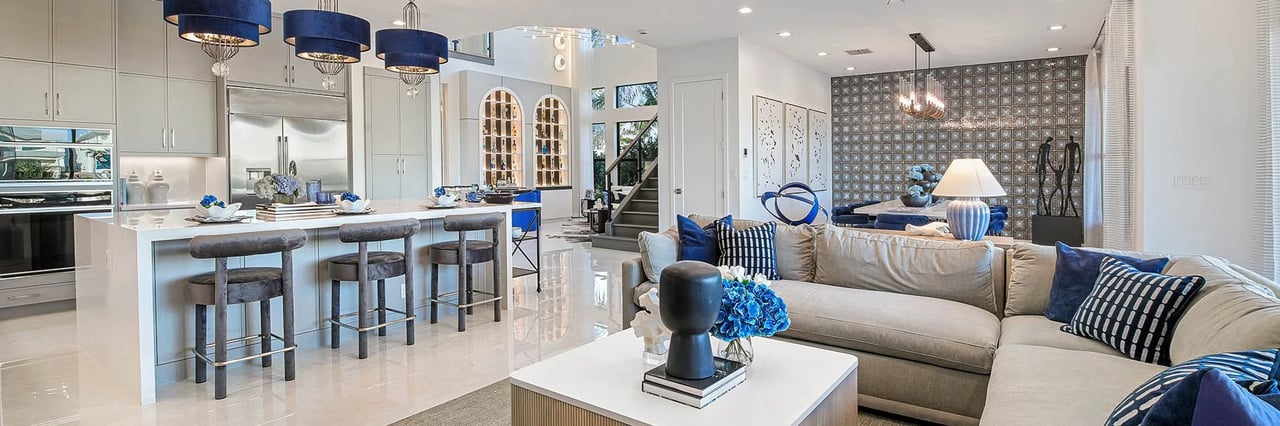Open-plan kitchen and living room with a white island, blue pendant lights, and a neutral sectional sofa.