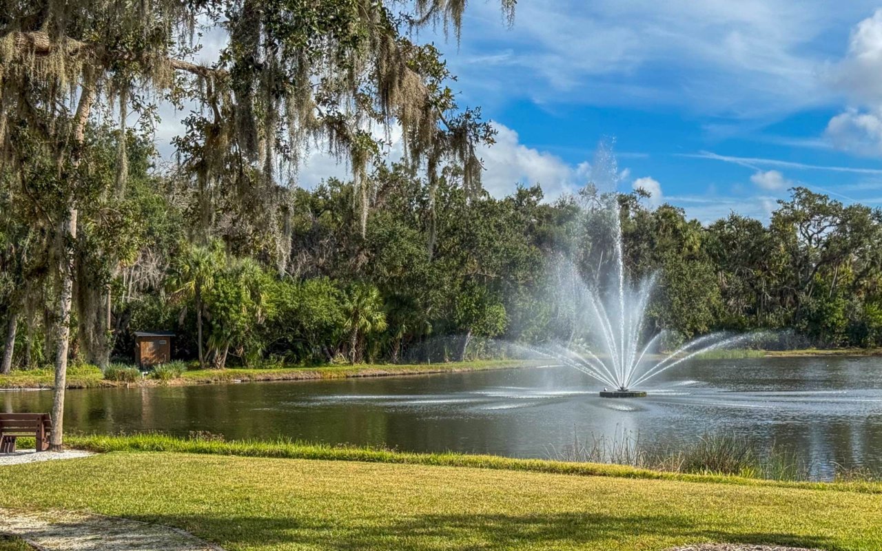 Community pond with a large fountain, moss-draped trees, grassy banks, and a bench.