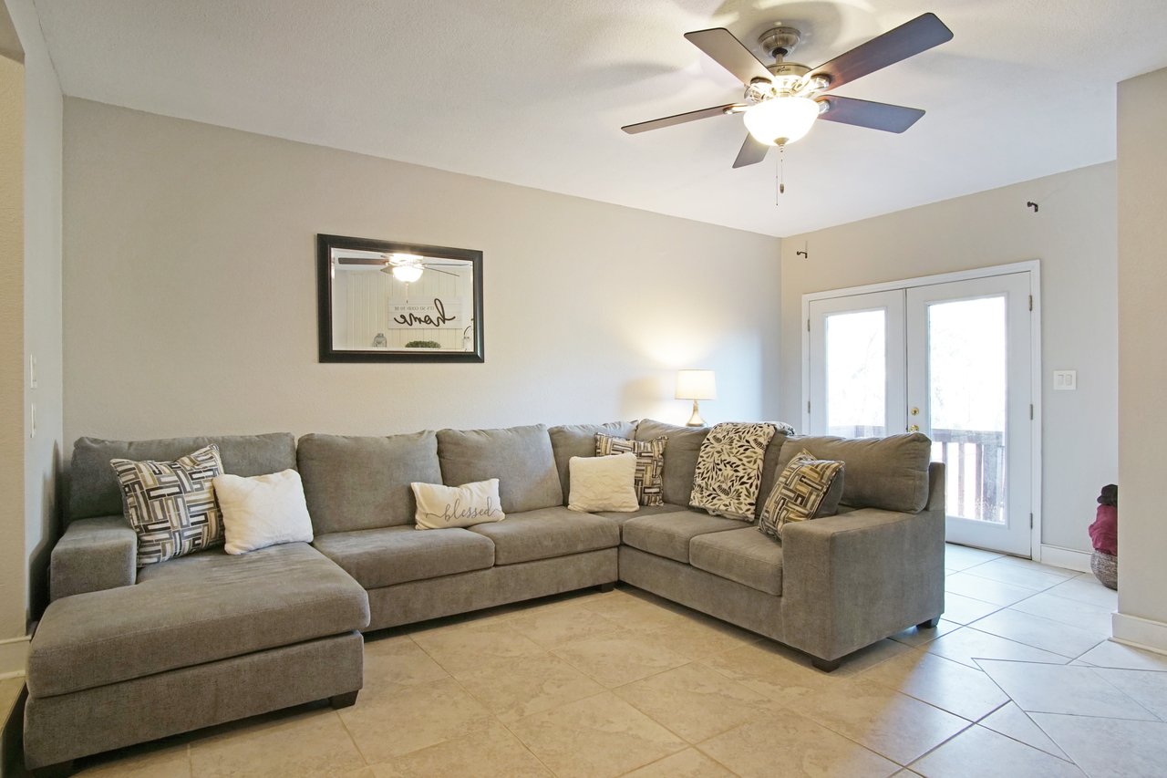 A cozy living room with a large gray L-shaped sofa adorned with patterned pillows and a "blessed" cushion. A mirror, ceiling fan, and lamp enhance the warm atmosphere.