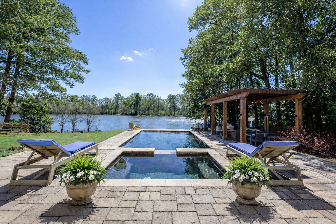 Backyard pool and stone patio overlooking a lake, with lounge chairs and a covered seating area.