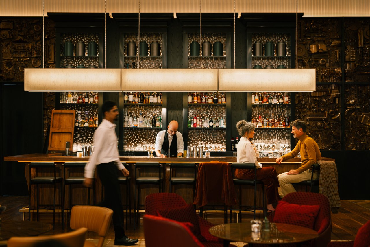 Bar interior with a long wood counter, illuminated bottle shelves, a bartender, and seated guests.