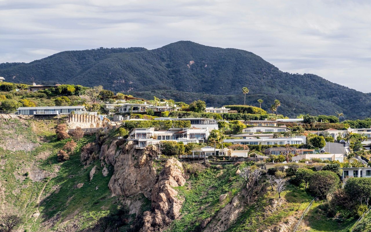 Hillside homes in Pacific Palisades overlook a rugged green slope beneath a forested mountain.