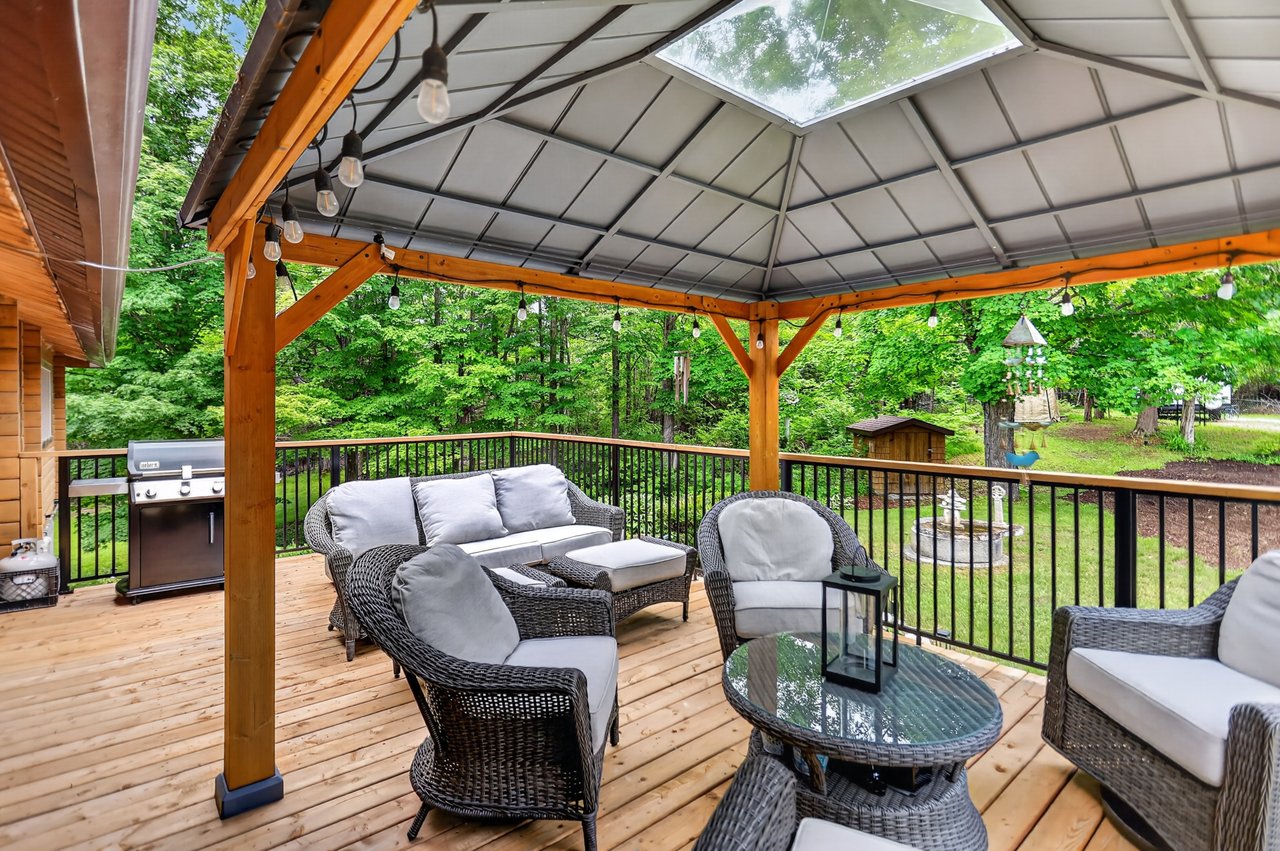 Covered wooden deck with wicker seating, skylight and barbecue beside a leafy yard.