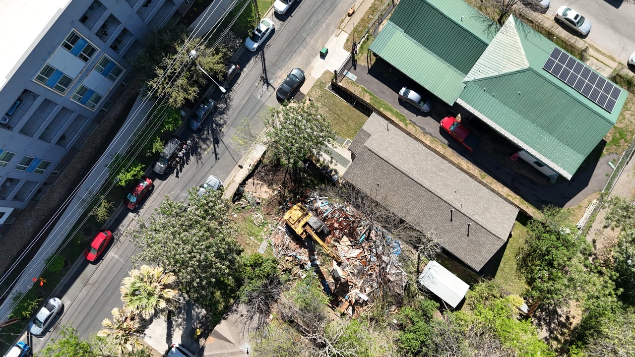 Aerial view of a demolition site with an excavator clearing rubble beside neighboring buildings.