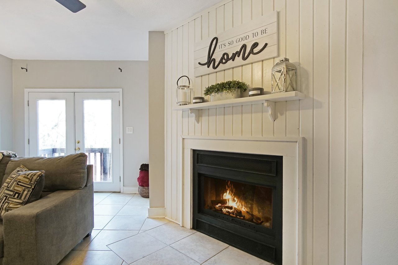 Cozy living room with a lit fireplace under a shelf with decor, including a "home" sign. A gray sofa and large windows create a welcoming atmosphere.