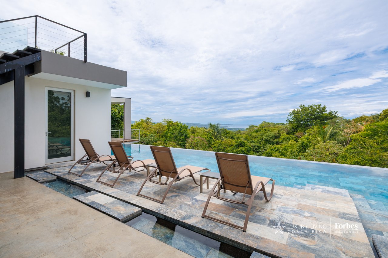 Infinity pool and stone terrace with loungers overlooking tropical forest and distant water.