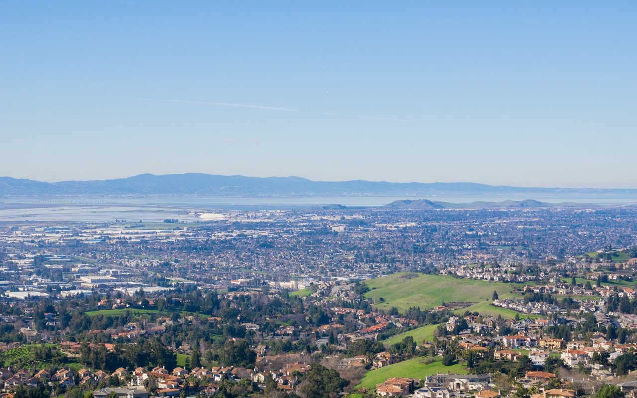 An expansive panoramic view of a vast urban area extending towards a large body of water in the distance, likely a bay, with hazy mountains beyond.